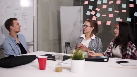 Group-Of-Three-Young-Coworkers-Sitting-In-The-Modern-Loft-Office-White-Huge-White-Table-Have-Fun-While-Break-Time,-Stick-Coloured-Papers-On-Forehead-And-Guessing-The-Word