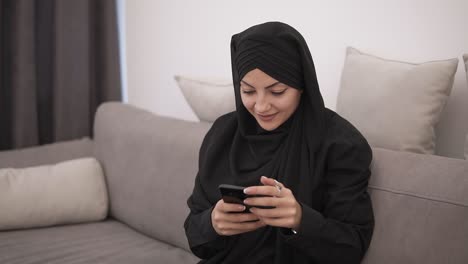 Beautiful-Muslim-Woman-In-Black-Hijab-Sitting-On-The-Sofa-And-Looking-To-Her-Smartphone-And-Smiling