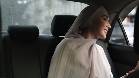 Portrait-Of-A-Young-Muslim-Woman-In-Beige-Headscarf,-Sitting-In-The-Car-While-Looking-Out-Through-The-Window-And-Smiling