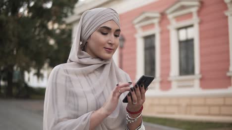 Muslim-Beautiful-Woman-In-Pastel-Hijab-With-Makeup-Standing-On-The-Street-Holding-Smartphone-While-Have-A-Walk-In-The-City