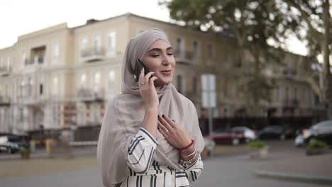 Muslim-Woman-In-Hijab-With-Makeup-Walking-On-The-Street-Talking-On-The-Phone-On-A-Background-Of-Beautiful-Old-Classic-Building