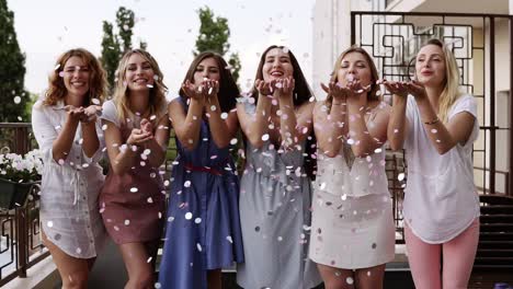 Front-View-Of-Six-Attractive-Young-Women-Playfully-Blowing-Bright-Colored-Confetti-From-Their-Hands-Together