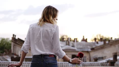 ,-Slender-Woman-In-Jeans-And-White-Shirt-Is-Standing-On-A-Balcony-With-Red-Flower-In-Her-Hand