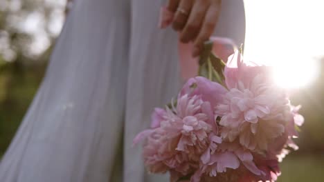 Backside-Tracking-Footage-Of-A-Woman-In-Bright-Clothes-Walking-By-The-Green-Meadow-With-Bouquet-Of-Pink-Flowers-In-Her-Hand
