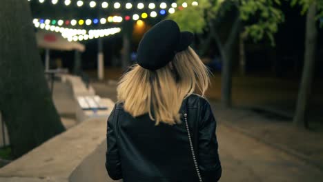 A-Beautiful-Girl-In-Stylish-Hat-And-A-Black-Leather-Jacket-Walks-Through-The-Night-Park-In-The-Light-Of-A-Cafe-Lamps