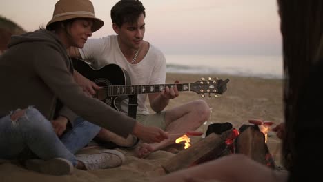 Der-Junge-Mann-Spielt-Gitarre-Am-Feuer-Und-Sitzt-Zusammen-Mit-Freunden-Am-Strand