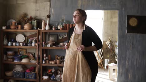 Slow-Motion-Footage-Of-Young-Woman-In-Casual-Clothes-In-Pottery-Workshop,-With-Ceramic-Wares-And-Supplies-On-Shelves-And-Table-With-Tools,-Putting-On-Beige-Apron-And-Smiling