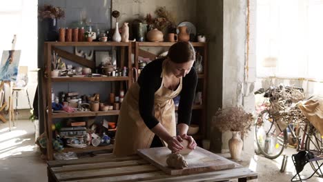 Portrait-Of-Female-Potter-Wearing-Beige-Apron-Putting-Clay-Piece-On-Worktop-And-Then-Starting-Kneading-It-With-Her-Hands