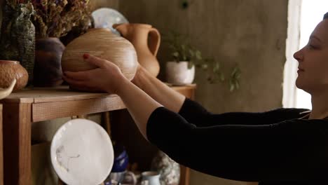 Side-View-Portrait-Of-Contemporary-Female-Potter-Putting-Handmade-Vase-On-Shelf-In-Studio,-Pleased-With-Her-Work,-Smiling-To-The-Camera