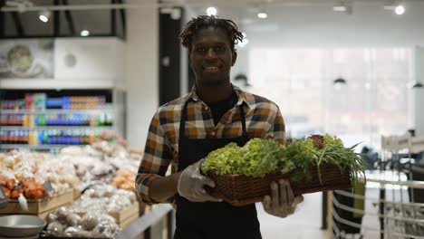 Male-Smiling-Worker-At-The-Store-Walks-With-Basket-Full-Of-Greens