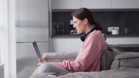 Woman-At-Home-Sitting-At-Kitchen-Sofa-Making-Video-Call-On-Laptop-Wearing-Headphones