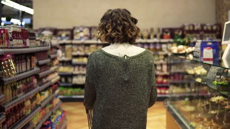 Rare-View-Of-A-Woman-With-Wavy-Hair-Is-Driving-Shopping-Trolley-Through-Food-Department-In-Supermarket-And-Looking-Around
