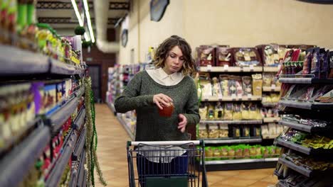 Front-View-Of-Woman-Walk-With-A-Cart-Near-Shop-Shelves-Choosing-A-Glass-Jar-In-Grocery-Market-And-Put-It-To-The-Cart