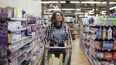 Modern-Young-Girl-In-Glasses-And-Headphones-On-Neck-Walking-With-Shopping-Cart-In-The-Supermarket-And-Stearing-Around
