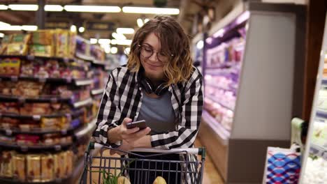 Woman-With-Smartphone-And-Headphones-On-Neck-Walking-With-Trolley-At-Supermarket-1