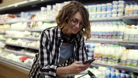Woman-With-Smartphone-And-Headphones-On-Neck-Walking-With-Trolley-At-Supermarket-2