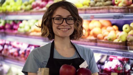 Beautiful-Smiling-Young-Female-Supermarket-Employee-In-Black-Apron-Holding-A-Box-Full-Of-Apples-In-Front-Of-Shelf-In-Supermarket-With-Pretty-Face-Looking-At-Camera-Professional-Front-Portrait-Startup-Business