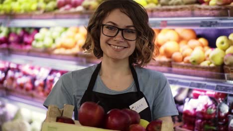 Beautiful-Smiling-Young-Female-Supermarket-Employee-In-Black-Apron-Holding-A-Box-Full-Of-Apples-In-Front-Of-Shelf-In-Supermarket-With-Pretty-Face-Looking-At-Camera-Professional-Front-Portrait-Startup-Business-1