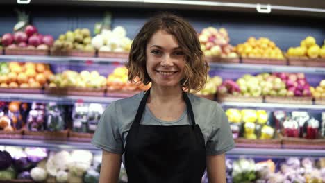 Portrait-Of-Attractive-Young-Saleswoman-In-Black-Apron-Standing-In-Supermarket-With-Shelves-Of-Fruits-On-Background,-Looking-At-Camera-And-Smiling