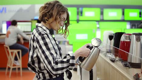 Side-View-Of-A-Young-Cheerful-Positive-Attractive-Woman-In-Plaid-Shirt-Choosing-Electronic-Kettle-In-Household-Appliances-Store,-Standing-In-A-Row,-Holding-And-Examines-Silver-Kettle