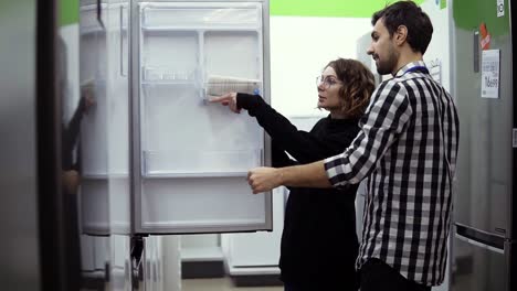 Junge-Ehepaare-Prüfen-Kühlschrank-Mit-Offener-Tür,-Design-Und-Qualität-Vor-Dem-Kauf-In-Einem-Unterhaltungselektronikgeschäft
