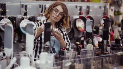 In-The-Appliances-Store,-An-Attractive-Curly-Woman-In-A-Plaid-Shirt-Chooses-A-Blender-Stick-By-Viewing-And-Holding-The-Device-In-Her-Hands