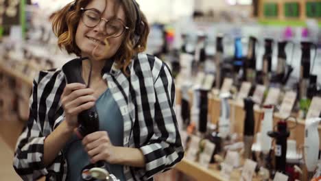 Portrait-Of-Cute-Pretty-Young-Woman-With-Short-Curly-Hair,-Plaid-Shirt-And-In-Headphones-Dancing-And-Pretending-She's-Singing-In-The-Middle-Of-The-Supermarket-With-Household-Using-Blender-Stick-As-Microphone