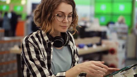 Curly-Woman-With-Headphones-On-Neck-Standing-At-The-Counter-With-Mobile-Phones-In-Casual-Clothes-Choosing-A-New-Smartphone-In-A-Modern-Electronics-Store