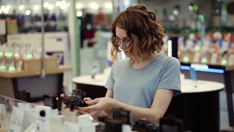 Young-Curly-Woman-In-Glasses-Shopping-For-A-New-Photocamera-In-The-Electronics-Store