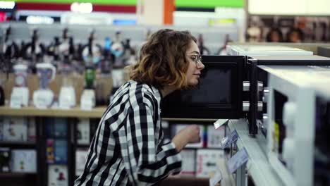 Side-View-Of-A-Young-Curly-Haired-Woman-In-Plaid-Shirt-Chooses-A-Microwave-Oven-In-A-Consumer-Electronics-Supermarket