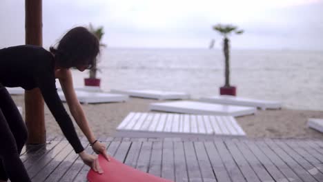 Woman-In-Black-Unfolding-A-Yoga-Mat-On-A-Wooden-Floor-Close-To-The-Water-On-The-Beach