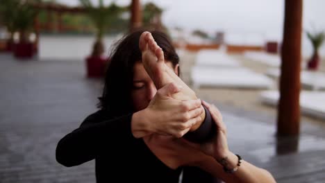 Mädchen-In-Schwarz-Macht-Yoga-Auf-Holzboden-Am-Strand,-Das-Mädchen-Führt-Yoga-Elemente-Durch,-Die-Das-Bein-Hinter-Der-Schulter-Beugen