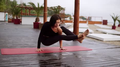 Mädchen-In-Schwarz-Macht-Yoga-Auf-Holzboden-Am-Strand,-Das-Mädchen-Führt-Yoga-Elemente-Durch,-Die-Das-Körpergewicht-Auf-Den-Händen-Ausgleichen