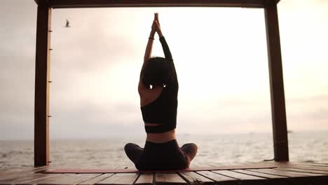 Mujer-Practicando-Yoga-Y-Estirando-La-Columna-Vertebral-Y-Las-Manos-Sentadas-En-Una-Alfombra-En-El-Muelle-Del-Mar-Inclinándose-A-Los-Lados,-Vista-Trasera