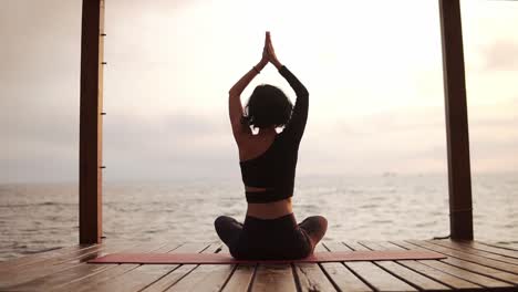 Mujer-Practicando-Yoga-Y-Estirando-La-Columna-Vertebral-Y-Las-Manos-Sentadas-En-Una-Alfombra-En-El-Muelle-Del-Mar-Tomándose-De-La-Mano-Por-Encima-De-La-Cabeza