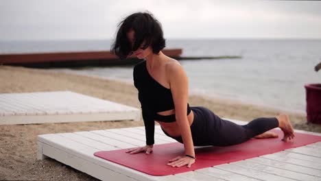 Mujer-Relajante-En-Ropa-Deportiva-Negra-Practicando-Yoga-En-La-Alfombra-Frente-Al-Mar-Por-La-Mañana