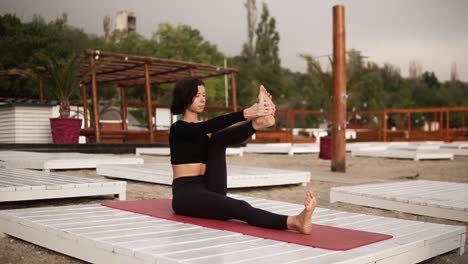Mujer-Practicando-Yoga-Sentada-En-Una-Alfombra-En-La-Playa-Realizando-Asanas-Y-Elementos-De-Yoga-O-Estirando-Las-Piernas
