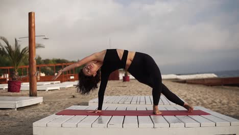 Frau,-Die-Yoga-Auf-Der-Matte-Am-Strand-Praktiziert-Und-Yoga-Asanas-Und--Elemente-Durchführt-Oder-Sich-Dehnt,-Den-Körper-Biegt