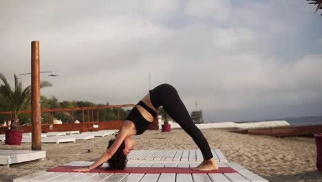Mujer-Practicando-Yoga-En-La-Alfombra-En-La-Playa-Realizando-Asanas-Y-Elementos-De-Yoga-O-Estirando-Las-Piernas-Levantándolas-Con-El-Cuerpo-Doblado