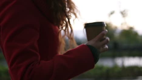 Junge-Frau-Im-Roten-Mantel-Trinkt-Kaffee-Auf-Der-Straße,-Während-Sie-An-Kalten-Herbsttagen-Spazieren-Geht