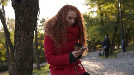 Woman-With-Curly-Long-Red-Hair-In-A-Red-Coat-And-Scarf-Standing-In-The-City-Park,-Using-Cell-Phone-And-Drinking-Coffee-From-Corton-Park
