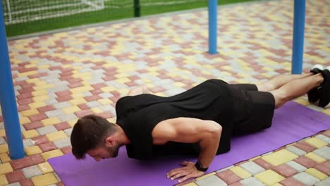 Man-In-Black-Sportswear-Doing-Push-Ups-On-Mat-With-Football-Field-On-Background-Outdoors