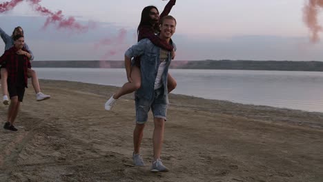 Friends-Having-Fun-Running-On-The-Beach-With-Smoke-Bombs,-Holding-Girls-On-Their-Backs