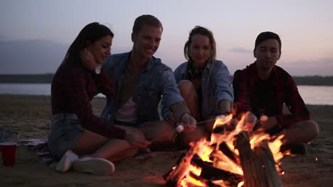 Young-And-Cheerful-Friends-Sitting-On-The-Wild-Beach-And-Fry-Marshmallows-Near-Bonfire