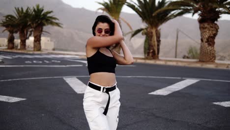 Stylish-Young-Woman-In-Sunglasses,-Black-Top-And-White-Pants-Dances-Funky-In-The-Middle-Of-The-Road-Slow-Motion-Woman-Freerly-Moves-Her-Body
