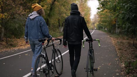 Dos-Amigos-Cercanos-Dando-Un-Agradable-Paseo-Con-Bicicletas-En-El-Parque-De-La-Ciudad-Entre-árboles-Altos-Verdes-Y-Amarillos,-Concepto-De-Estilo-De-Vida-Activo,-Comunicación,-Citas