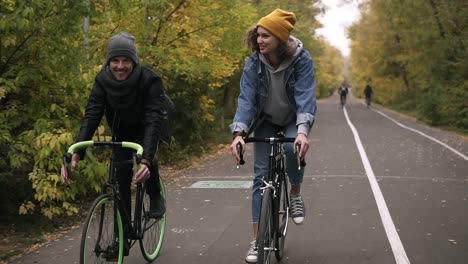 Amigos-Jóvenes-Y-Sonrientes-O-Una-Pareja-Joven-Con-Sombreros-En-Bicicleta-En-Sus-Bicicletas-De-Trekking-Por-El-Parque-De-Otoño-En-Bicicleta