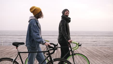 Side-View-Of-Smiling-Couple-Of-Young-Hipsters-Walking-Together-With-Their-Bikes-Near-The-Sea-At-Autumn-Day