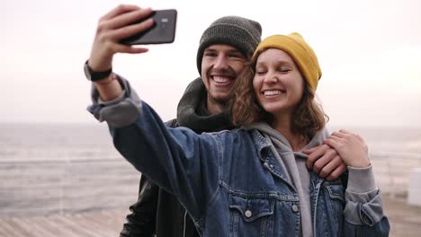 Happy-Young-Couple-Taking-Selfie-By-Mobile-Phone-Near-The-Seaside-In-Cloudy-Day