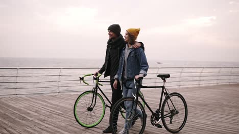 Beautiful-Smiling-Couple-Of-Young-Hipsters-Walking-Together-Embracing-With-Their-Bikes-Near-The-Sea-At-Autumn-Day